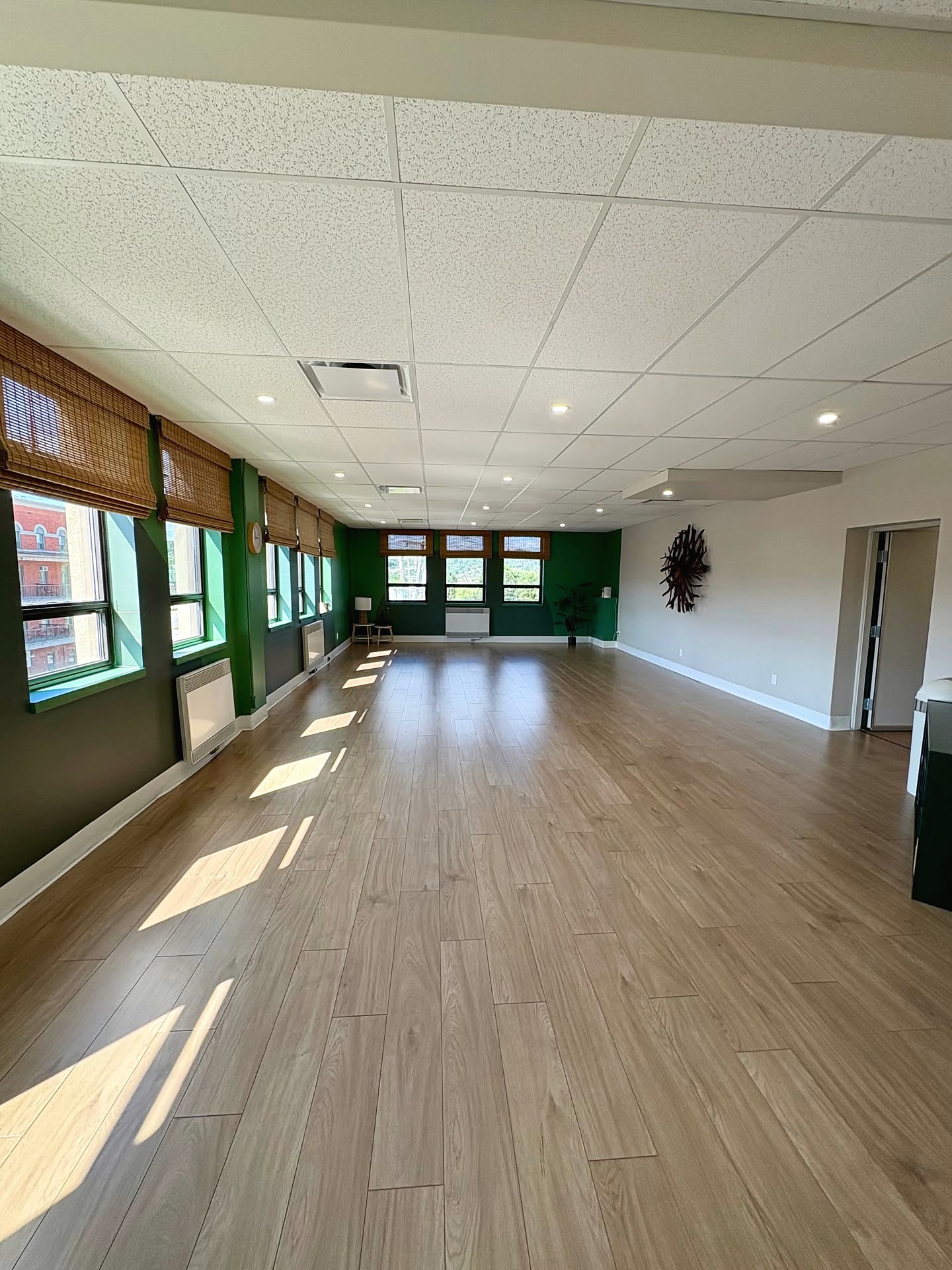 Grande salle de pratique vide au Centre Bodhi avec fenêtres, lumière naturelle et plancher de bois.